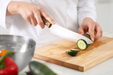 Professional chef cutting fresh cucumber at white table in kitchen, closeup