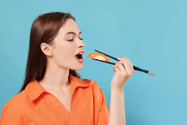 Beautiful young woman eating sushi with chopsticks on light blue background