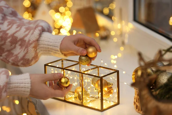 Christmas atmosphere. Woman putting beautiful baubles into decorative container on window sill indoors, closeup