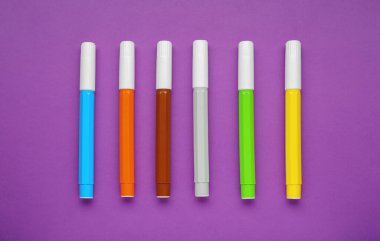 Different colorful markers on purple background, flat lay
