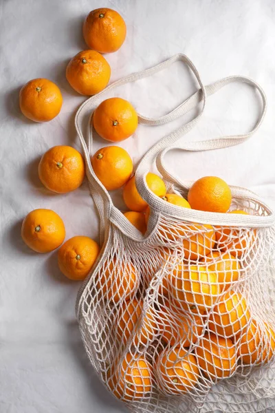 Net bag with many fresh ripe tangerines on white cloth, flat lay