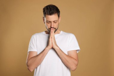 Ellerini kenetleyip bej arka planda dua eden bir adam.