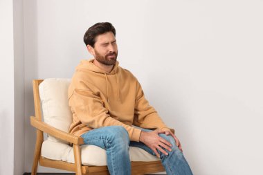 Man suffering from leg pain and touching knee on soft armchair indoors, space for text