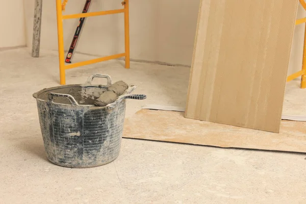 Bucket of adhesive mix with spatula near tile on floor indoors