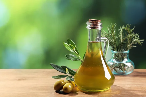Jug of cooking oil, olives and green leaves on wooden table against blurred background. Space for text