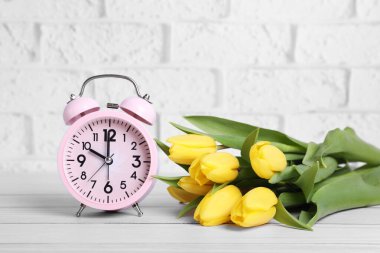 Pink alarm clock and beautiful tulips on white wooden table against brick wall. Spring time
