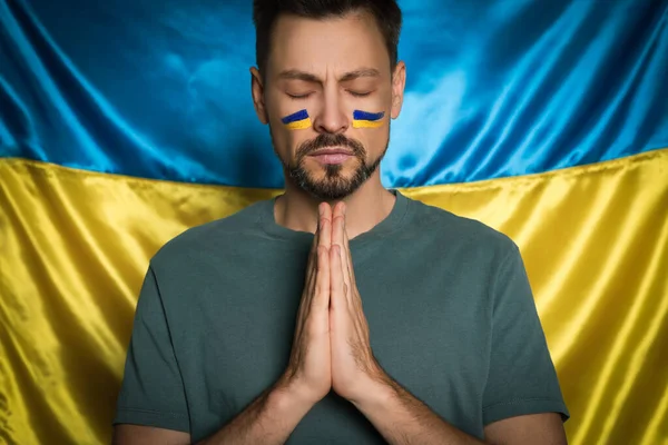 Man with clasped hands praying near Ukrainian flag