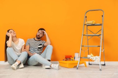 Tired designers sitting on floor with painting equipment near freshly painted orange wall indoors