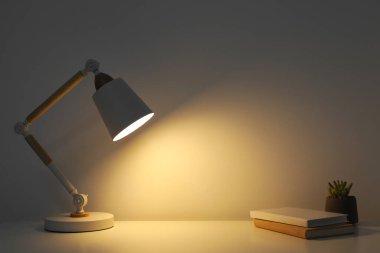 Stylish modern desk lamp, books and plant on table near white wall indoors