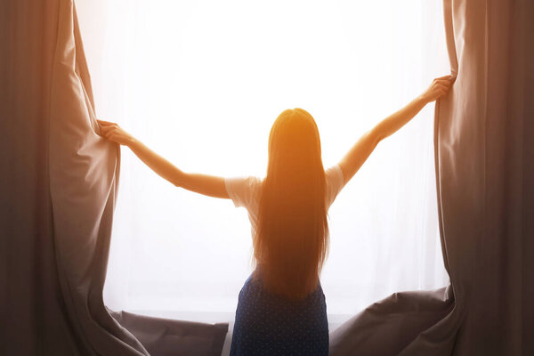 Woman opening stylish curtains at home, back view