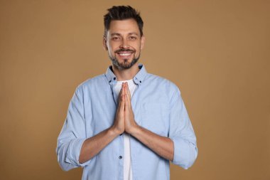 Ellerini kenetleyip bej arka planda dua eden bir adam.