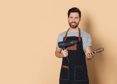 Smiling hairdresser in apron holding dryer and brush on light brown background, space for text
