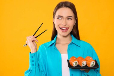 Happy young woman with plate of sushi rolls and chopsticks on orange background