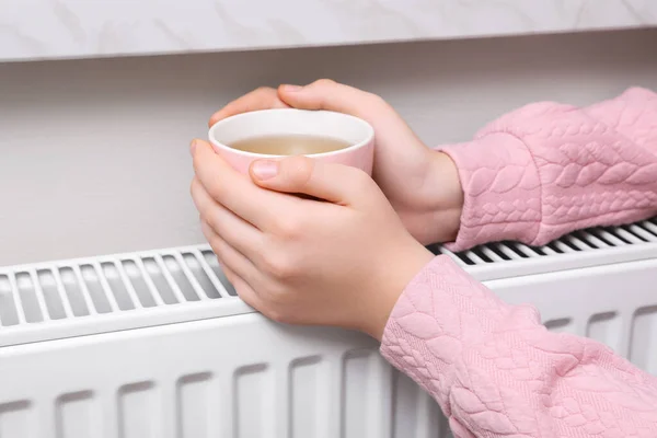 Girl with cup of tea warming hands on heating radiator indoors, closeup