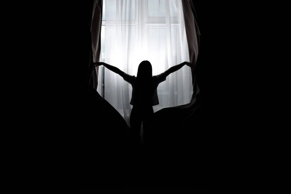 Silhouette of woman opening curtains at home, back view