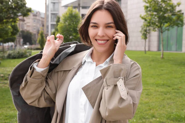 Woman holding garment cover with clothes while talking on phone outdoors. Dry-cleaning service