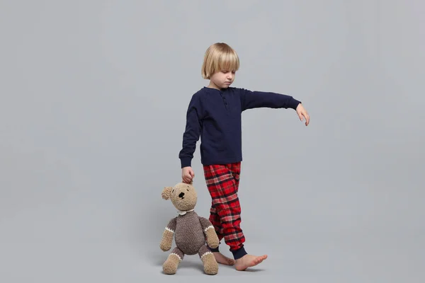 Boy in pajamas with toy bear sleepwalking on light gray background