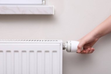 Girl adjusting heating radiator thermostat near white wall indoors, closeup