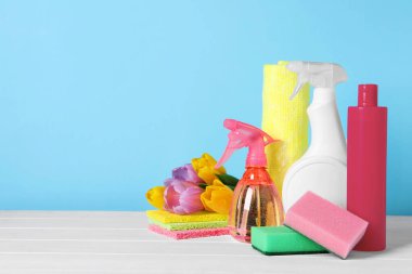 Spring cleaning. Detergents, flowers and sponges on white wooden table against light blue background. Space for text