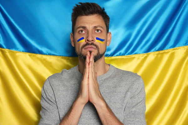 Sad man with clasped hands praying near Ukrainian flag