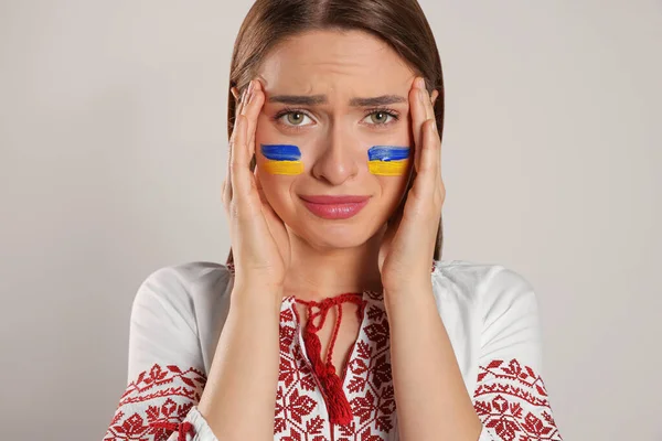 Sad young woman with drawings of Ukrainian flag on face against beige background