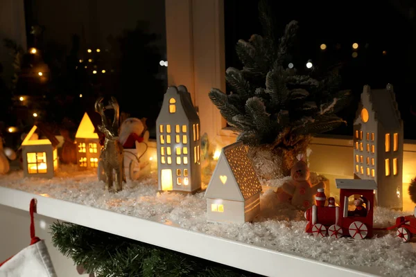 Christmas atmosphere. Beautiful glowing houses, fir trees, artificial snow and toys on window sill indoors