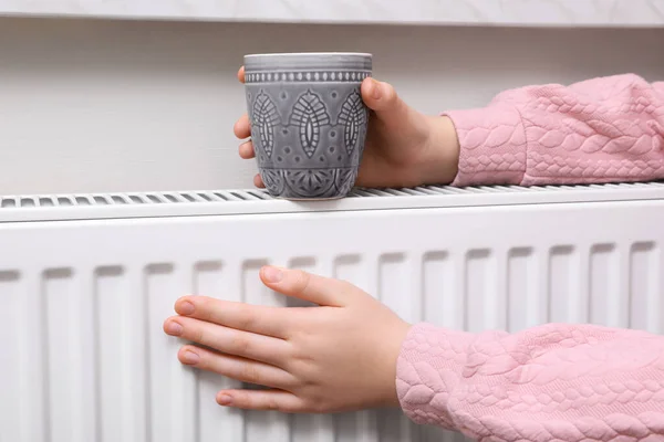 Girl with cup of drink warming hands on heating radiator indoors, closeup