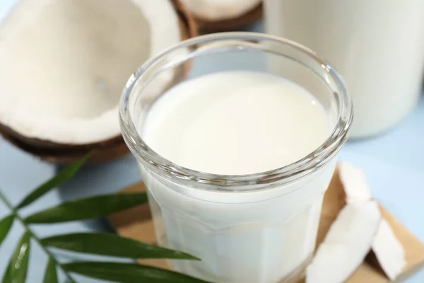 Glass of delicious vegan milk, coconuts and leaf on light blue background, closeup