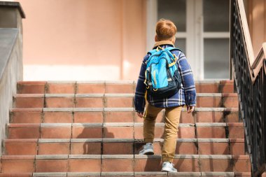 Little boy with backpack going up stairs to school, back view