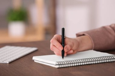 Woman writing in notebook at wooden table indoors, closeup. Space for text