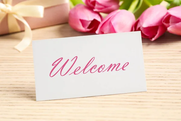 Welcome card, gift box and beautiful pink tulips on wooden table, closeup