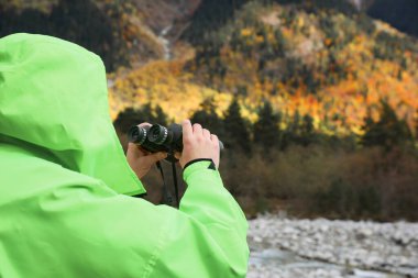 Woman holding binoculars near river in beautiful mountains, closeup. Space for text