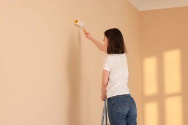 Young woman painting wall with roller on stepladder indoors, space for text. Room renovation