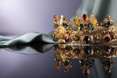 Beautiful golden crown with gems near light cloth on dark mirror surface. Space for text