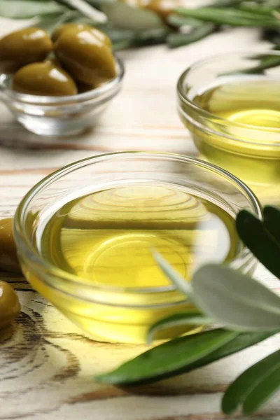 Bowls with cooking oil, olives and green leaves on white wooden table, closeup