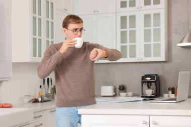 Emotional young man checking time while drinking coffee in kitchen. Being late