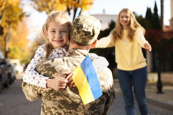 Daughter hugging her father in military uniform and holding Ukrainian flag on city street. Family reunion
