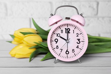 Pink alarm clock and beautiful tulips on white wooden table against brick wall, closeup. Spring time