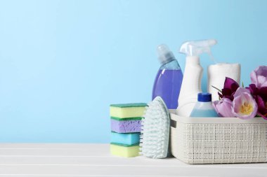 Spring cleaning. Basket with detergents, flowers and tools on white wooden table against light blue background. Space for text