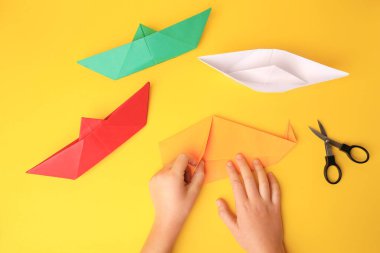 Origami art. Child folding paper on yellow background, closeup and top view