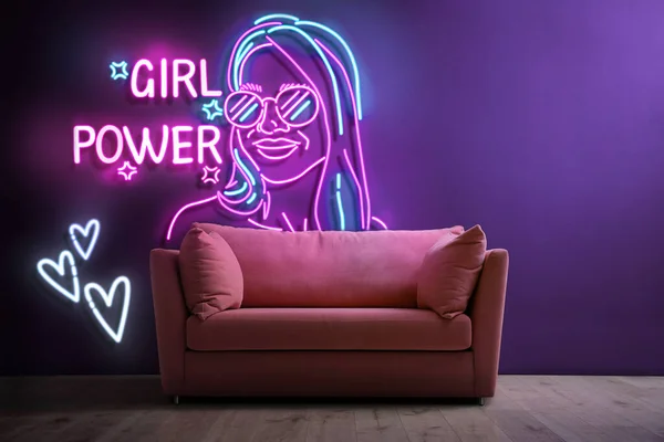Glowing neon sign with outline of woman, hearts and words Girl Power near couch on wall indoors