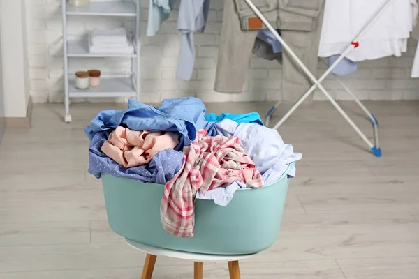 Plastic laundry basket overfilled with clothes on white stool indoors