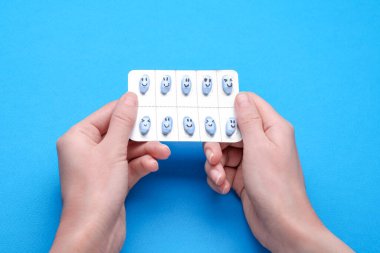 Woman holding antidepressants with different emoticons on light blue background, top view