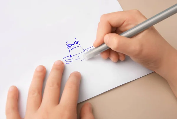Child erasing drawing with erasable pen on paper sheet against beige background, closeup