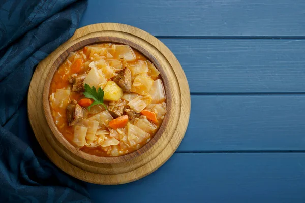 Tasty cabbage soup with meat, carrot and parsley on blue wooden table, top view. Space for text