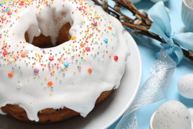 Delicious Easter cake decorated with sprinkles near painted eggs and willow branches on light blue background, closeup