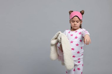 Girl in pajamas and sleep mask with toy bunny sleepwalking on gray background, space for text clipart