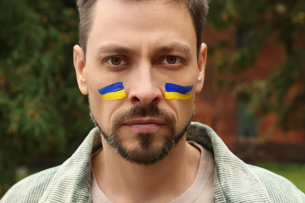 Angry man with drawings of Ukrainian flag on face outdoors, closeup