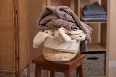 Wicker laundry basket overfilled with clothes on wooden stool indoors