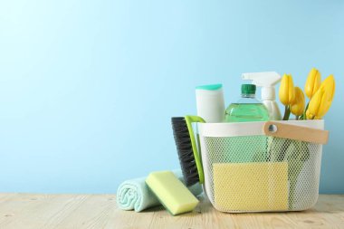 Basket with different cleaning supplies and beautiful spring flowers on wooden table against light blue background. Space for text